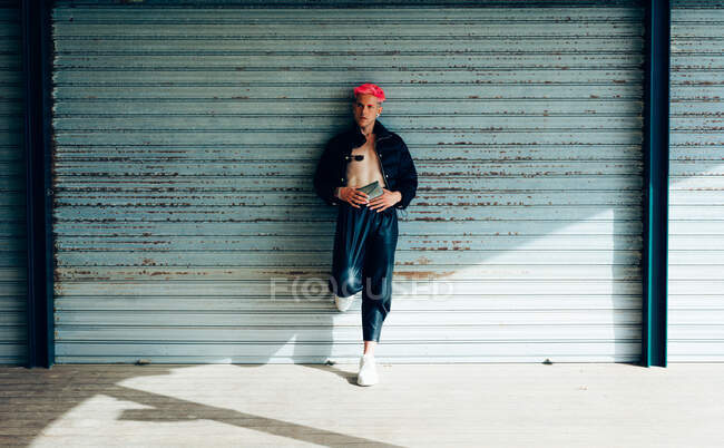 Homme transsexuel en tenue tendance avec sac à main et cheveux roses regardant loin contre le mur altéré par une journée ensoleillée — Photo de stock