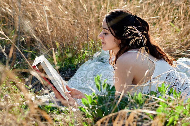 Мечтательная очаровательная брюнетка в белом платье, лежащая на полевом лугу и читающая книгу при солнечном свете — стоковое фото