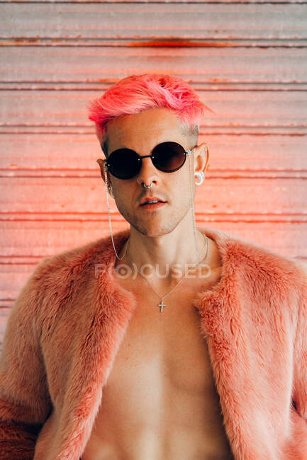 Giovane gay in occhiali da sole moderni e giacca di pelliccia elegante con capelli rosa contro muro grigio — Foto stock