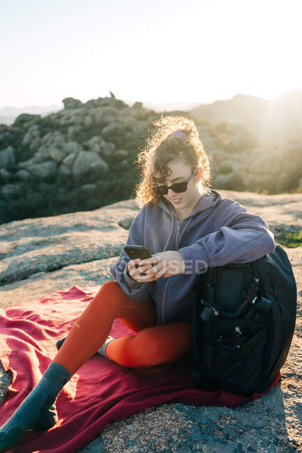 Joyful young female hiker with curly hair in casual outfit and sunglasses leaning on backpack and using smartphone while relaxing on rocky hill slope on sunny day — Stock Photo