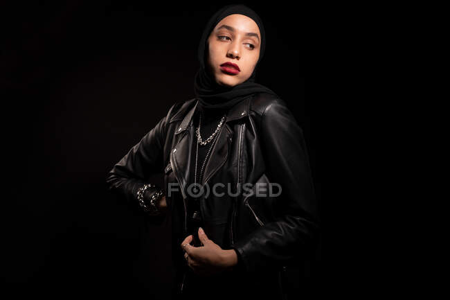 Attractive young Islamic female wearing black outfit with leather jacket and hijab gently looking away on black studio — Stock Photo