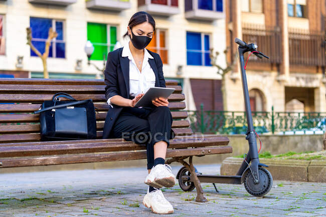 Etnica donna d'affari in panno maschera surf internet su tablet mentre seduto con le gambe incrociate su panchina urbana contro scooter — Foto stock