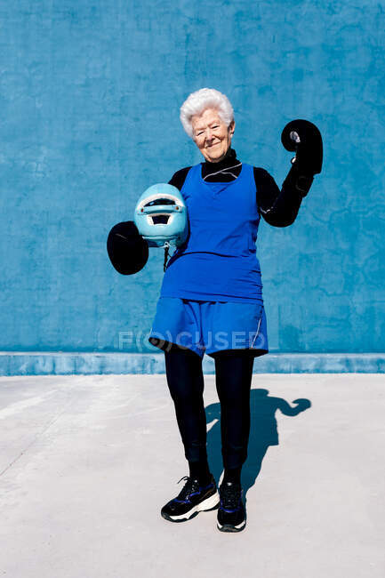 Повна довжина усміхнена зріла жінка в спортивному одязі і боксерських рукавичках, що стоять з шоломом в руці проти синьої стіни і дивиться на камеру — стокове фото