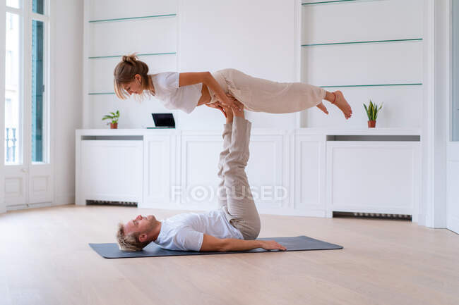 Side view of boyfriend lifting girlfriend while doing acro yoga together at home and holding hands — Fotografia de Stock