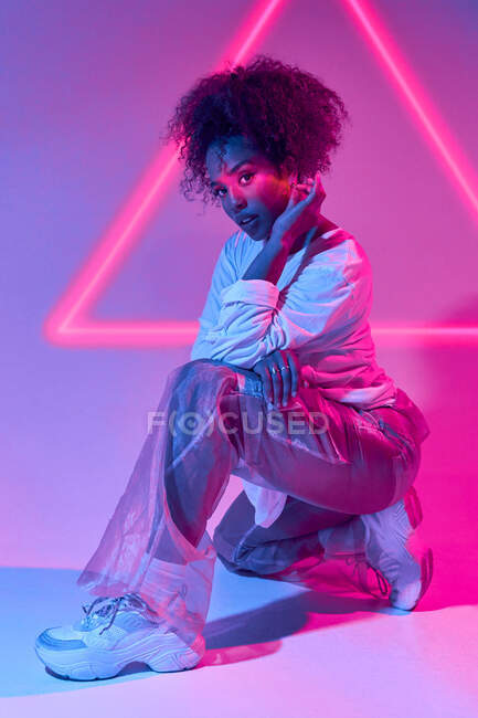 Fiducioso giovane donna afroamericana con i capelli ricci accovacciati guardando la fotocamera in luci al neon — Foto stock