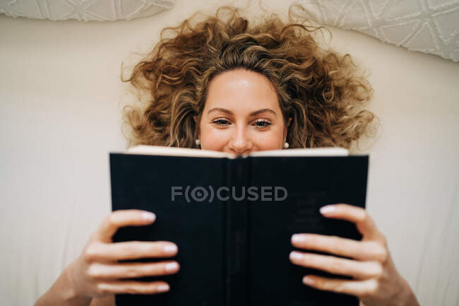Desde arriba feliz joven hembra con el pelo rizado acostado en la cama libro de lectura durante el fin de semana en casa - foto de stock