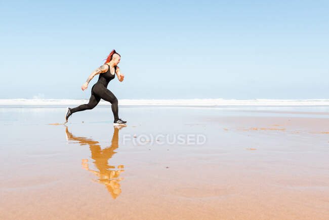 Seitenansicht einer aktiven Joggerin mit Tätowierungen, die am Meeresufer läuft, während sie während des Trainings im Wasser reflektiert — Stockfoto