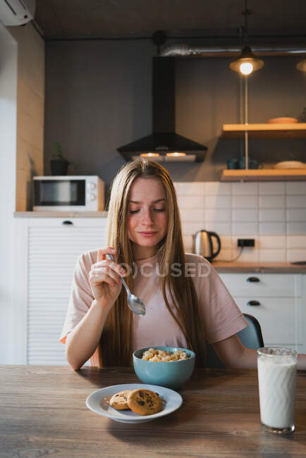 Joven hembra con cuchara y bol disfrutando de sabrosos anillos de maíz en la cocina - foto de stock