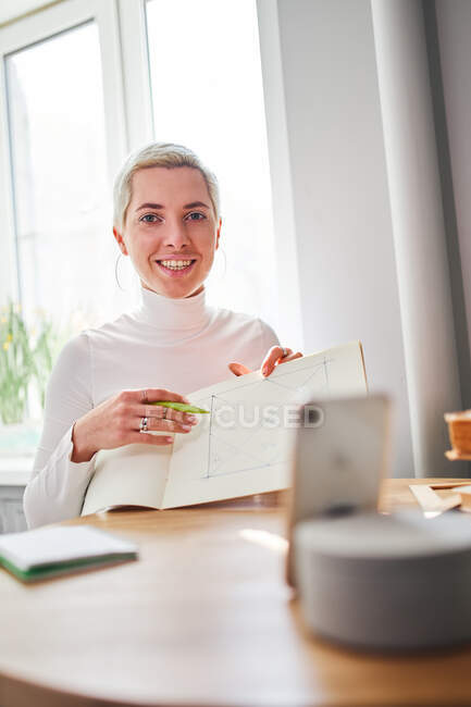 Улыбающаяся женщина-астролог демонстрирует геометрический рисунок в бумажном альбоме, глядя на камеру в доме в солнечный день — стоковое фото