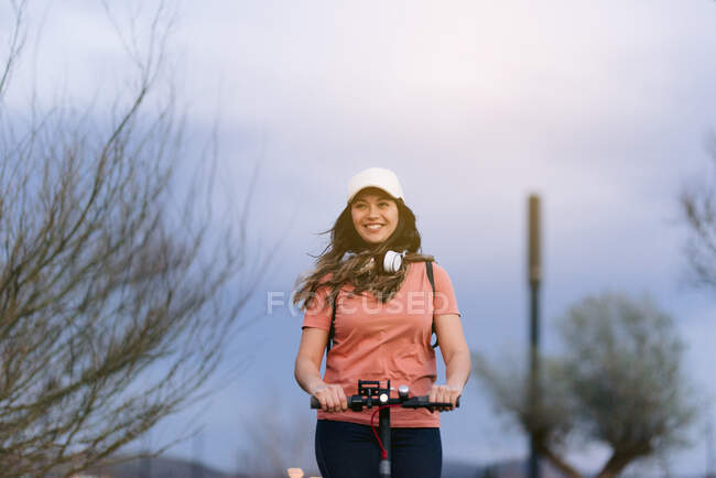 Mulher sorridente em boné com fone de ouvido em scooter elétrico olhando para longe sob céu nublado na cidade — Fotografia de Stock