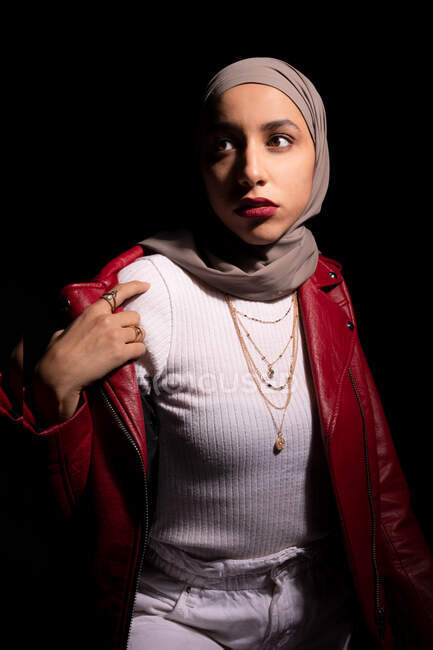 Confiant modish islamique femme portant foulard tenue tendance debout regardant loin sur fond noir en studio — Photo de stock