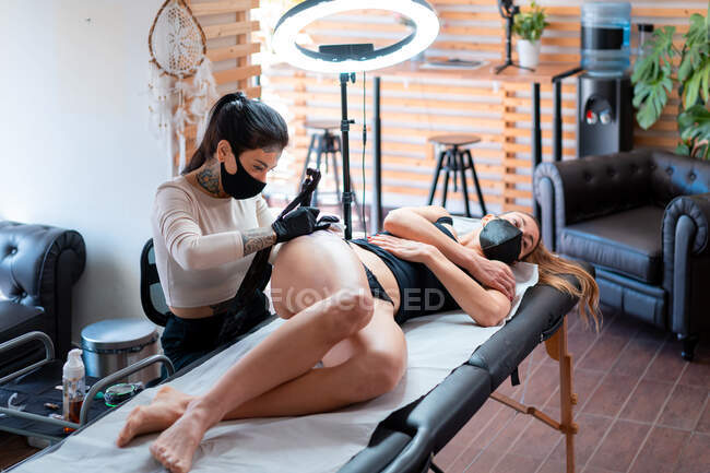 Жінка-татуювач в рукавичках з професійною машиною, що застосовує татуювання на тілі жінки в салоні — стокове фото
