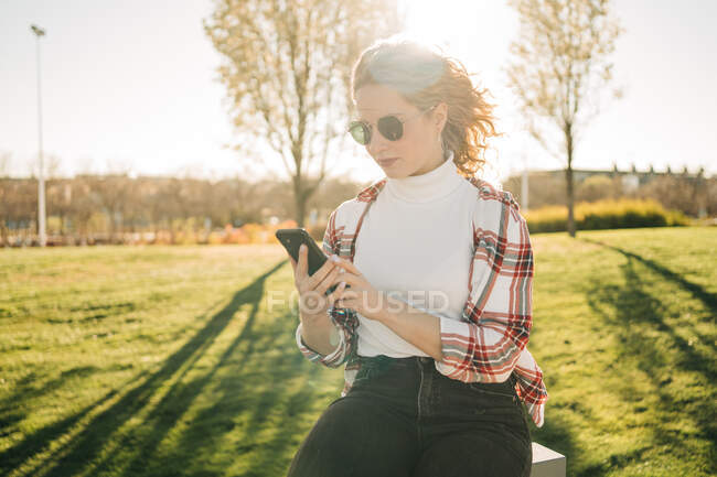 Trendy blonde female with curly hair sitting in a bench on green meadow in a park texting on mobile phone — Stock Photo