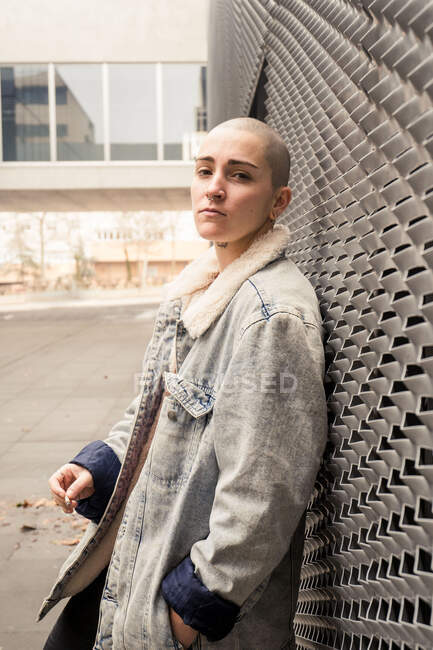 Vista lateral de una joven transexual segura de sí misma en chaqueta de mezclilla fumando cigarrillo mientras mira a la cámara - foto de stock