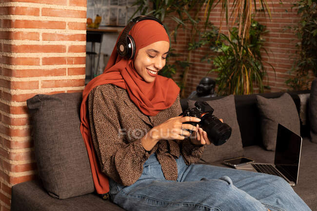 Jovem muçulmana encantada em uso modesto hijab e fones de ouvido usando câmera de foto profissional no sofá acolhedor — Fotografia de Stock