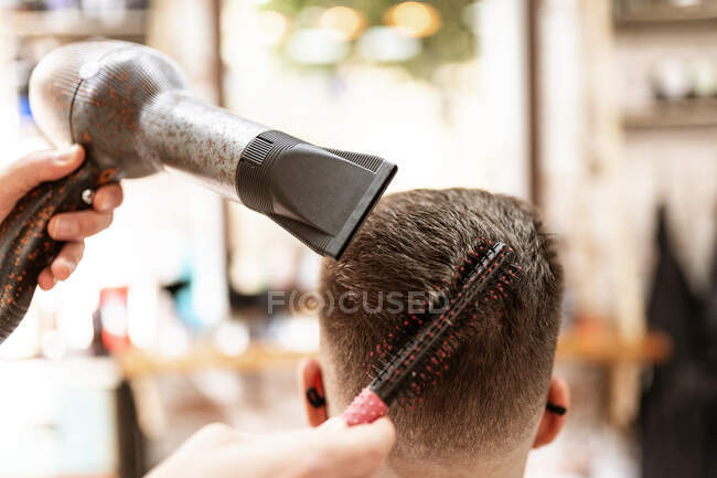 Estilista anónimo con secador de pelo contra hombre en capa en sillón en barbería - foto de stock