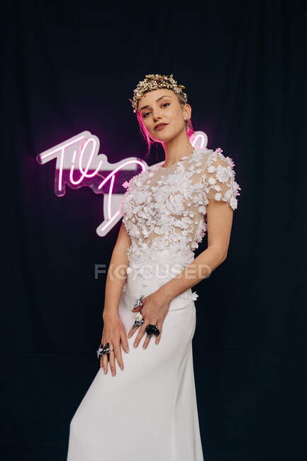 Charming young female model in bohemian elegant white lace gown and floral wreath standing against black background with neon inscription — Photo de stock