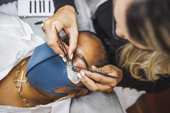 High angle of crop unrecognizable cosmetologist with tweezers applying fake eyelashes for extension on eye of ethnic client with face protective mask in salon during coronavirus pandemic — Fotografia de Stock