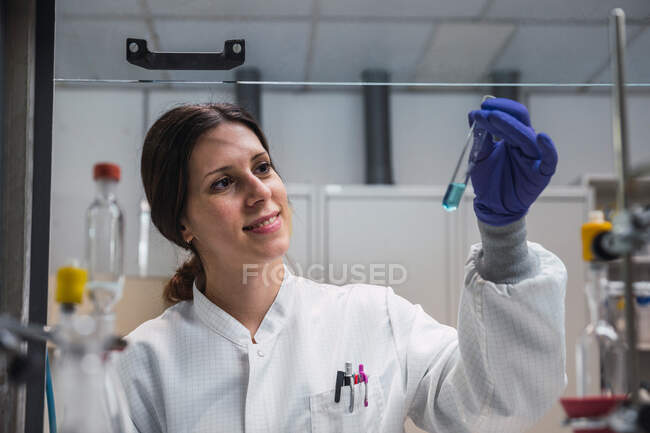 Cheerful professional female scientist in protective gloves and robe looking at blue solution n test tube while working in equipped laboratory — Stock Photo