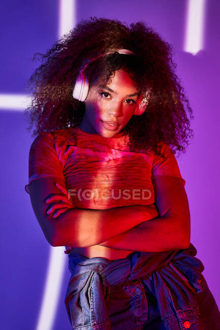 Splendida giovane donna afroamericana in abbigliamento casual ascoltare musica in cuffia mentre in piedi con le braccia incrociate in luci al neon e guardando la fotocamera — Foto stock