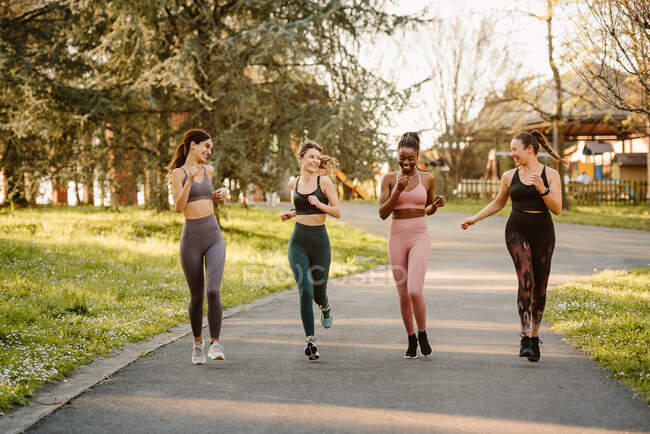 Smiling multiracial female runners in activewear jogging and talking during cardio training on walkway in town — Stock Photo