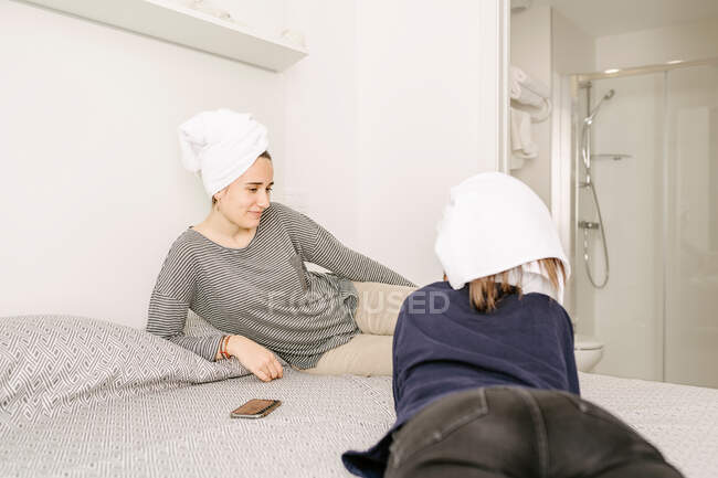 Happy young ethnic female best friends in casual clothes and towels on heads relaxing on comfortable bed after having bath at home — Stock Photo