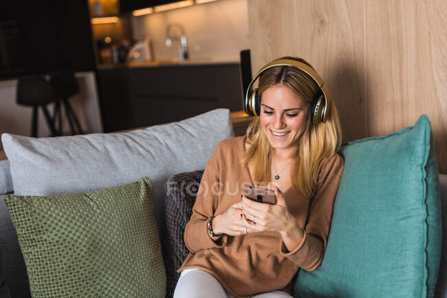 Fröhliche Frau sitzt auf dem Sofa und genießt Musik im Kopfhörer, während sie auf den Bildschirm ihres Smartphones schaut — Stockfoto