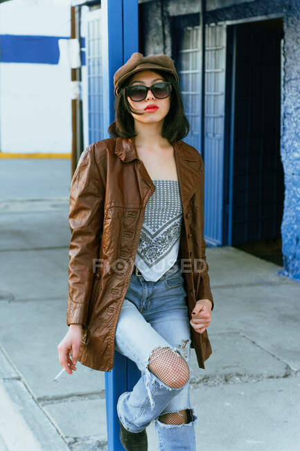 Молодая этническая женщина в кожаной куртке и солнцезащитных очках, стоящая с рукой на бедре и глядя на улицу — стоковое фото