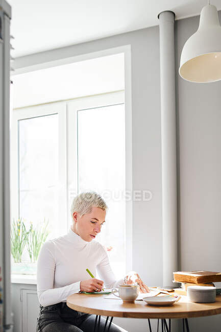 Astrologista feminina tomando notas no bloco de notas na mesa com xícara de café em casa à luz do sol — Fotografia de Stock