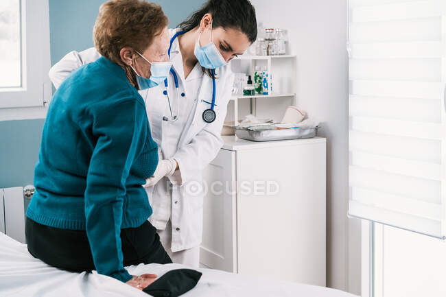 Обрезание женщины-врача в одноразовой маске и униформе помогает пожилой женщине встать во время обследования в больнице во время пандемии коронавируса — стоковое фото