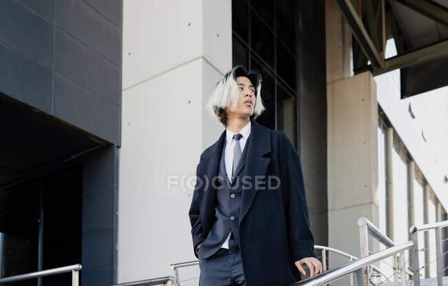 Jovem Asiático executivo masculino de terno com as mãos nos bolsos olhando para longe entre corrimãos na cidade — Fotografia de Stock