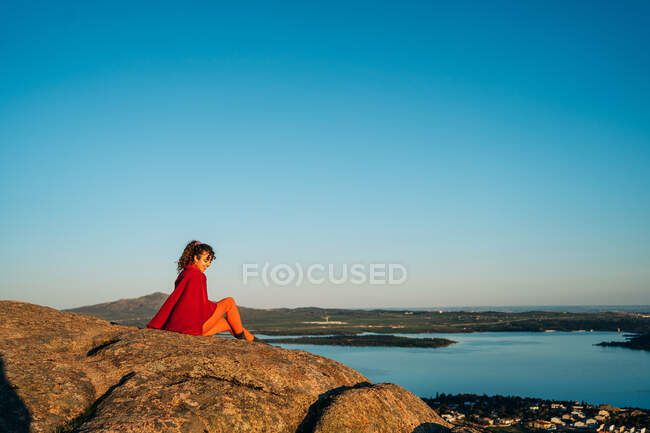 Vue latérale d'une jeune voyageuse enchantée aux longs cheveux foncés en tenue décontractée assise sur une falaise rocheuse et admirant la mer contre un ciel sans nuages au coucher du soleil — Photo de stock