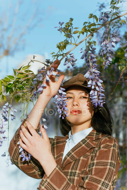 Young gentle ethnic female in checkered wear with beret touching blossoming plant while looking away on street — Foto stock