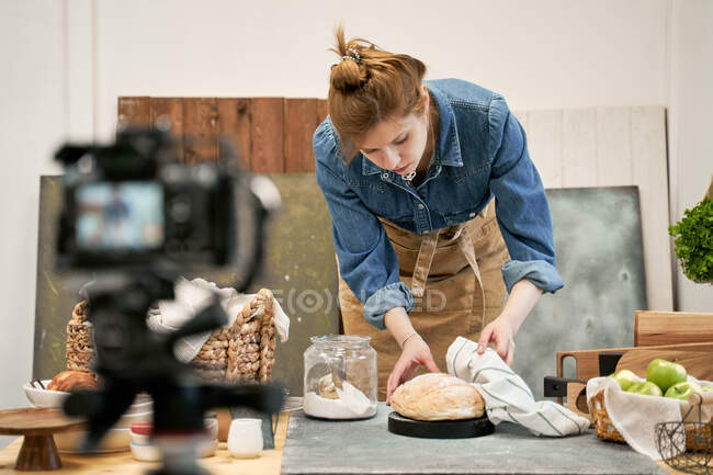 Jovem blogueira feminina com pão de trigo fresco contra a câmera fotográfica durante o processo de cozimento à mesa em casa — Fotografia de Stock