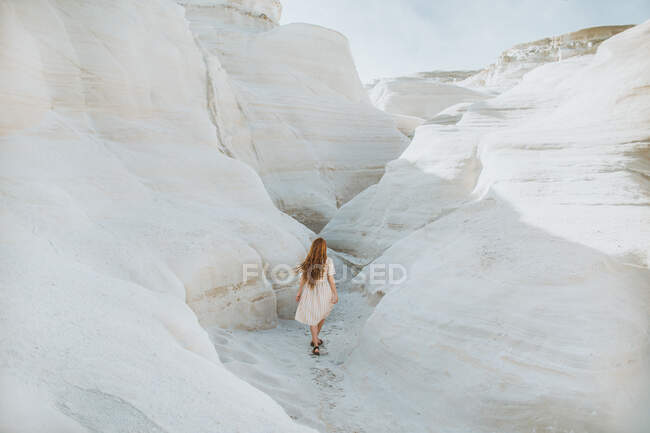 Vue de dos femelle anonyme en robe de soleil blanche se promenant le long d'un passage étroit formé par de légers rochers courbés par temps ensoleillé à Sarakiniko Grèce — Photo de stock