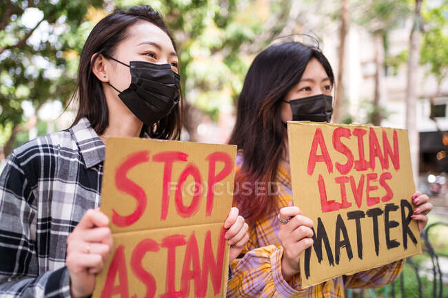 Mujeres étnicas con máscaras sosteniendo carteles protestando contra el racismo en la calle de la ciudad y mirando hacia otro lado - foto de stock