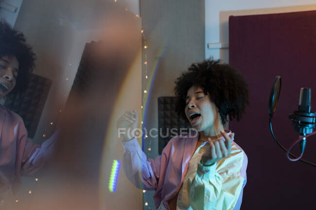 Black female singer performing song against microphone with pop filter while standing with hand on hip and closed eyes in sound studio — Stock Photo