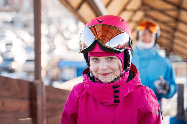 Menina alegre vestindo capacete de esqui rosa e roupas esportivas quentes em pé no ensolarado clube de esportes ao ar livre e olhando para a câmera com sorriso — Fotografia de Stock