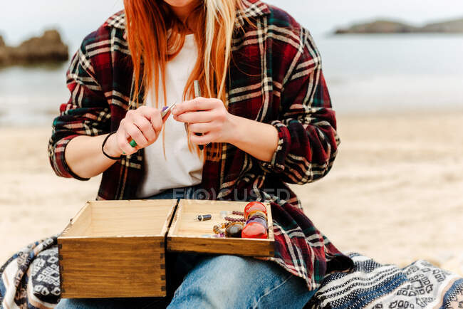 Cultivez une artisan anonyme créant une bijouterie artisanale tout en utilisant des pierres semi-précieuses et assis sur la plage près de la mer — Photo de stock
