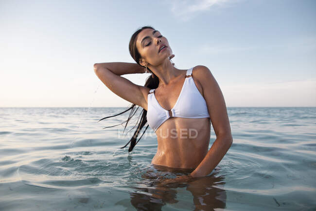 Mujer femenina joven en traje de baño blanco tocando el cabello mojado mientras mira hacia otro lado en el mar ondulado - foto de stock