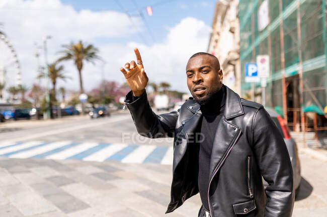 Trendy African American male wearing black leather jacket raising hand to catch taxi while standing on sunny city street — Photo de stock