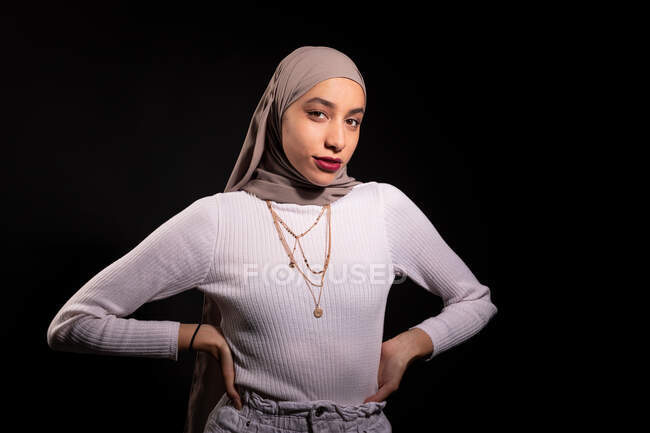 Modish fiducioso femminile musulmana in hijab in piedi e guardando la fotocamera in studio scuro — Foto stock