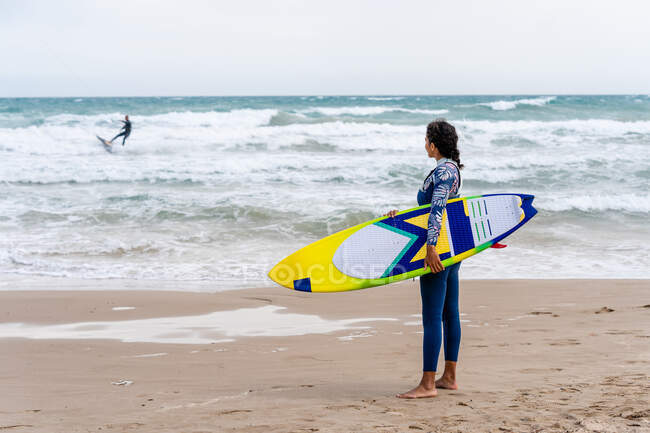 Atleta feminina étnica alegre em wetsuit com kiteboard em pé na costa arenosa contra pipa inflável — Fotografia de Stock