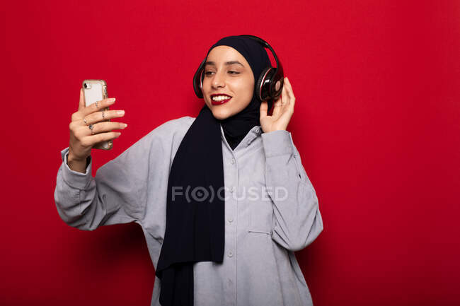 Alegre mujer islámica vistiendo atuendo casual y hijab escuchando buena música en auriculares inalámbricos y navegando por teléfono inteligente sobre fondo rojo en el estudio - foto de stock