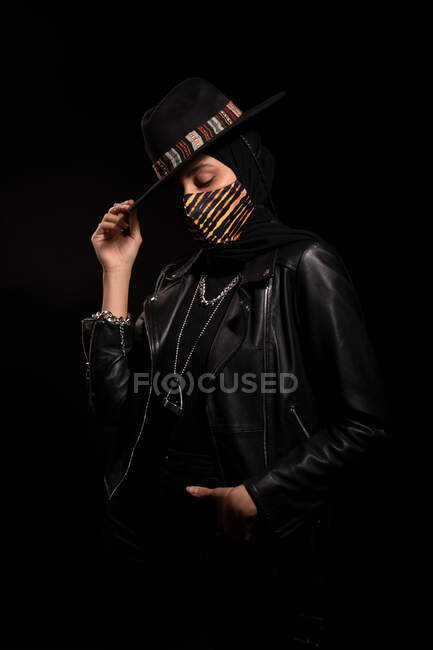 Fashionable Islamic female wearing leather jacket hijab and trendy hat standing with hand in pocket and touching brim in dark studio during coronavirus outbreak — Stock Photo