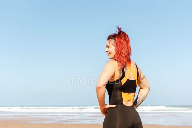 Rückenansicht einer selbstbewussten Athletin mit den Händen auf den Hüften, die am Sandstrand des Ozeans im Sonnenlicht wegschaut — Stockfoto