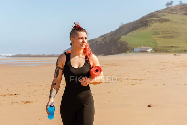 Atleta sonriente con alfombra enrollada y botella de agua paseando por la costa arenosa del mar mientras mira hacia otro lado - foto de stock