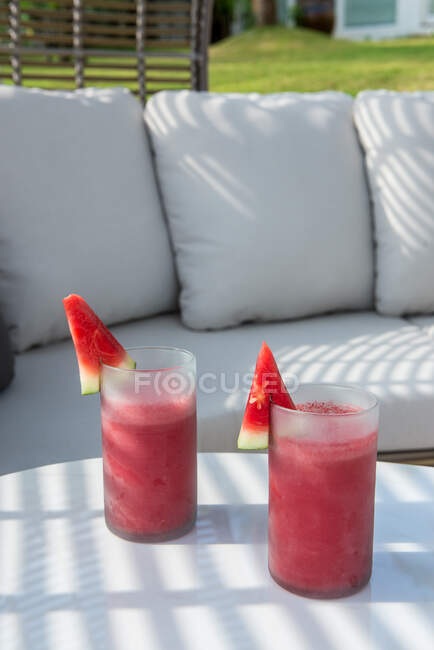 High angle pair of glasses of fresh squeezed watermelon smoothie served on table near soft comfy sofa on grassy lawn in garden — Photo de stock