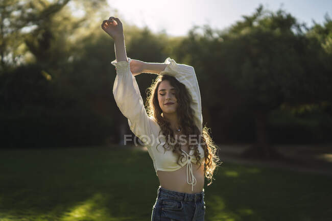 Stylish young female with bare belly standing on lush grassy lawn in sunny park and closed eyes — Stock Photo