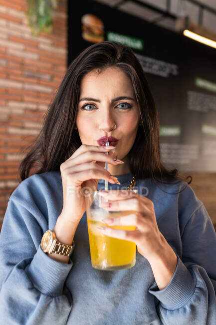Веселая молодая женщина в голубом свитере, потягивающая холодный газированный безалкогольный напиток через солому, проводя свободное время в кафетерии и счастливо глядя в камеру — стоковое фото